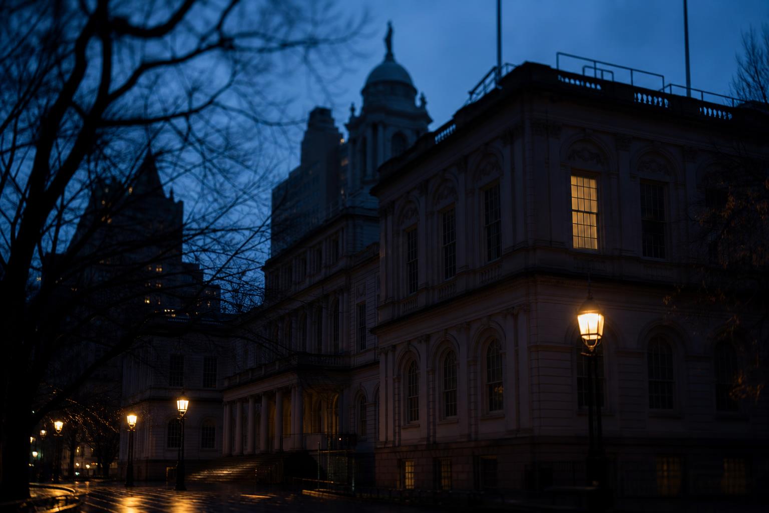New York City Hall facade at deep dusk with a single warm window glowing on the dark stone, bare tree branches silhouetted in the foreground and wet pavement reflecting amber streetlights — the building where Mamdani's approval rating is being judged.